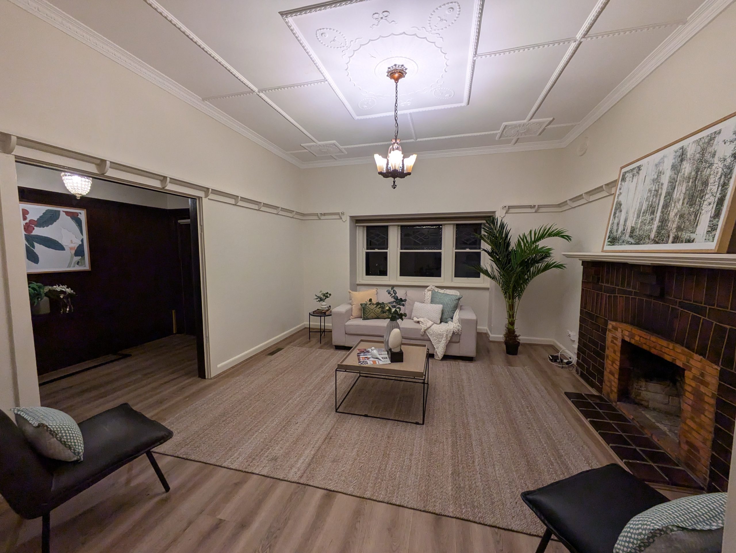 A cozy living room with a vintage ceiling design, a brick fireplace, and modern furniture. The room is softly lit, featuring a beige sofa, two black chairs, and a coffee table. A large plant and framed artwork decorate the space.