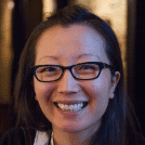A smiling woman with long dark hair wearing black glasses.